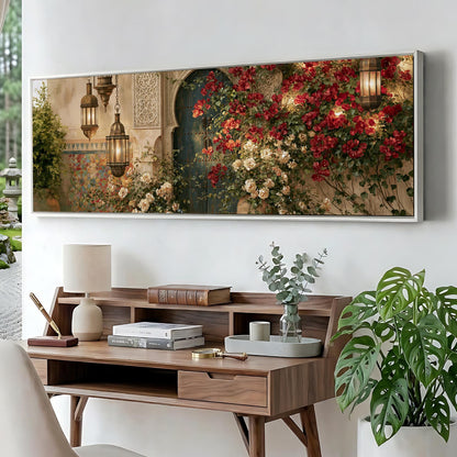 a large, framed painting of a garden scene with red flowers and greenery, hanging on a wall above a wooden desk with a lamp, books, and a vase of flowers.