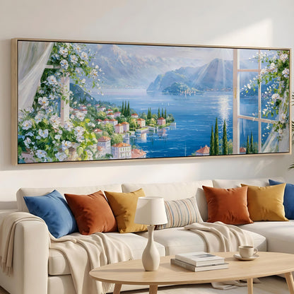 a large painting of a scenic view of a coastal town with a lake, mountains, and colorful flowers in the foreground. The painting is hung on a wall above a white sofa with colorful throw pillows.