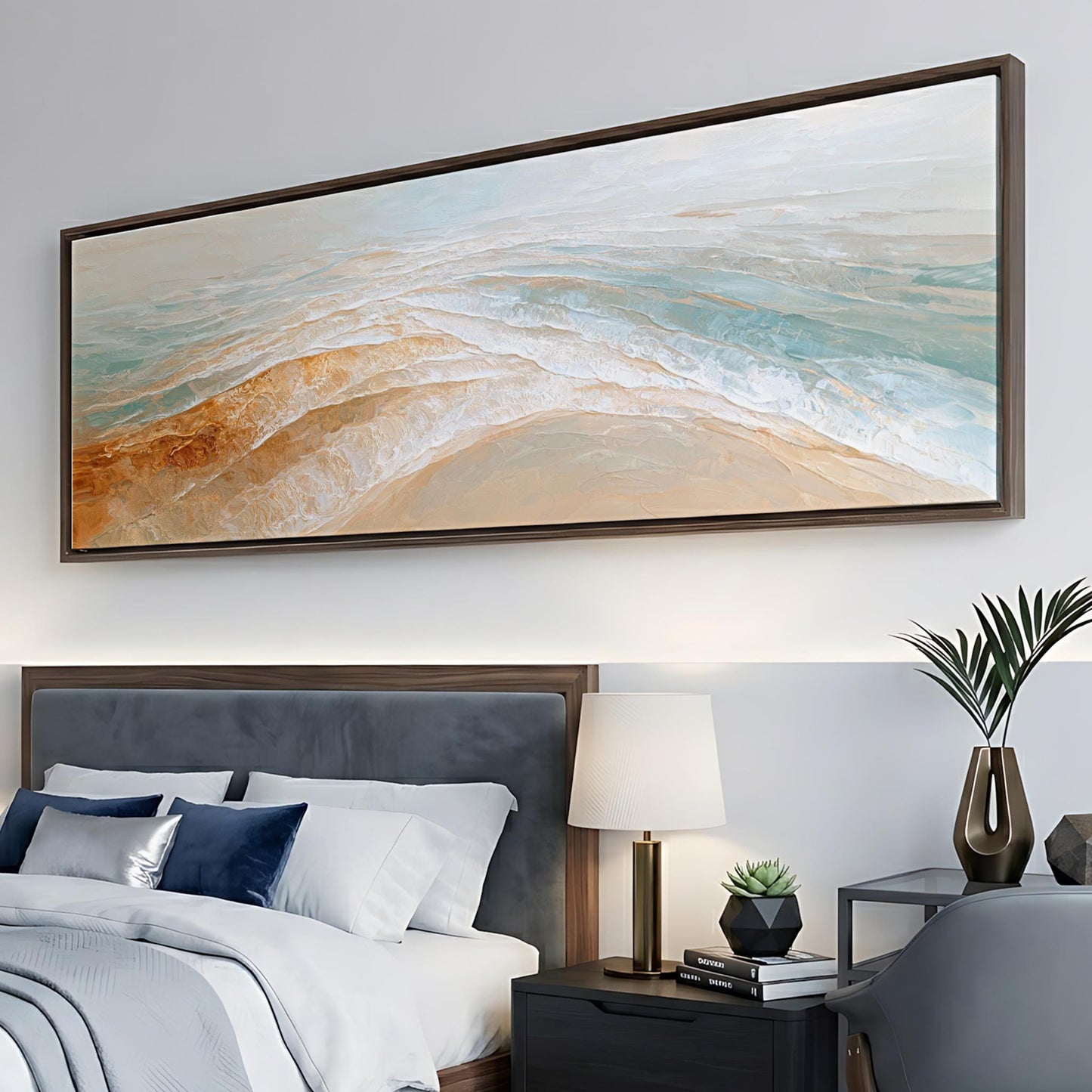a large, framed painting of a beach scene hanging above a bed with a gray headboard. The painting depicts a serene ocean landscape with waves gently lapping against the shore. The bed is adorned with white linens and a blue throw pillow, and a nightstand with a lamp and a plant is situated next to the bed.