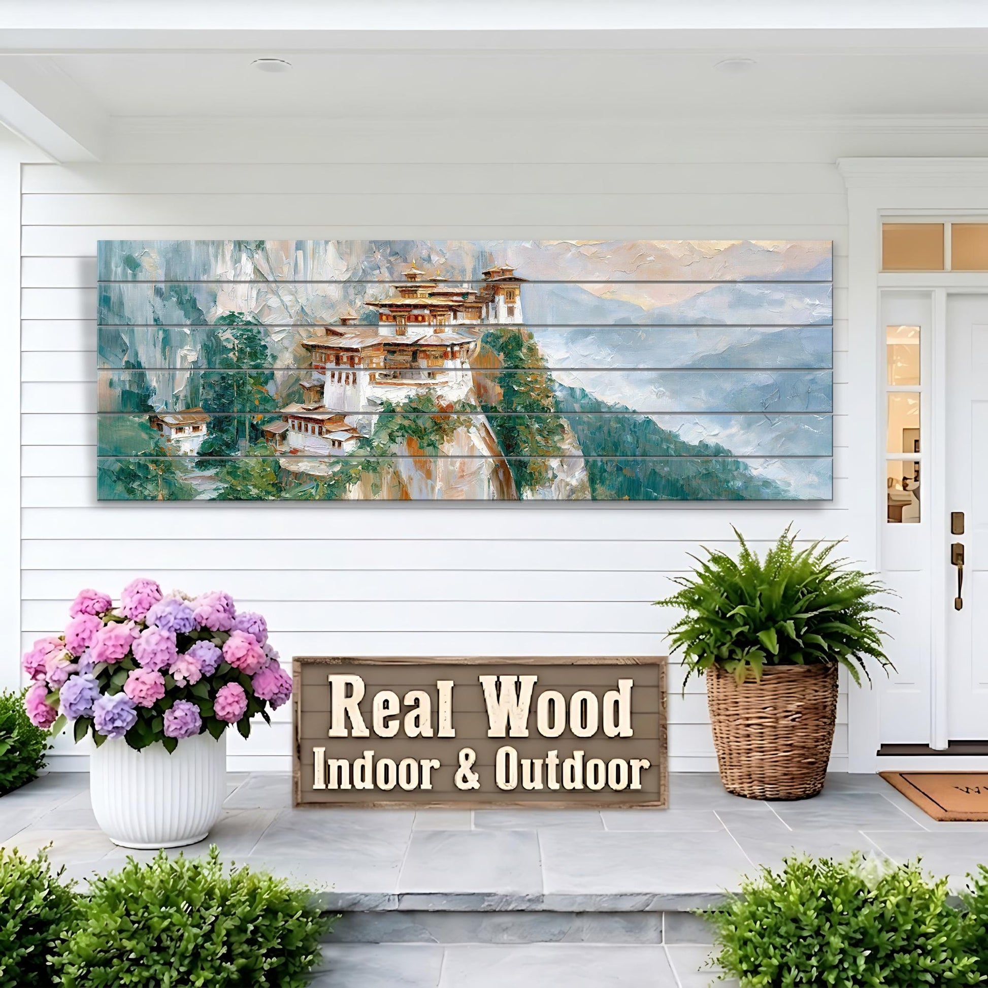 a large wooden sign that reads "Real Wood Indoor & Outdoor" in front of a white house with a painting of a mountain village on the wall. The house has a porch with potted plants and a wicker basket.