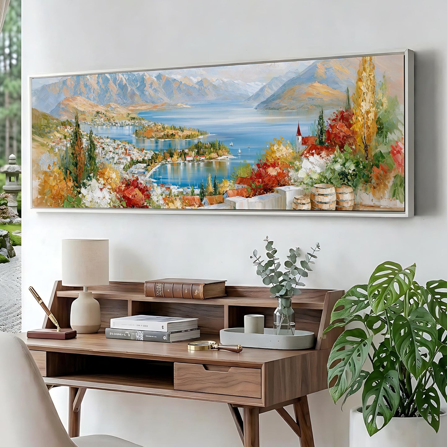 a large painting of a scenic landscape with mountains, a lake, and colorful foliage, hanging on a wall above a wooden desk with a lamp, books, and a vase of greenery.