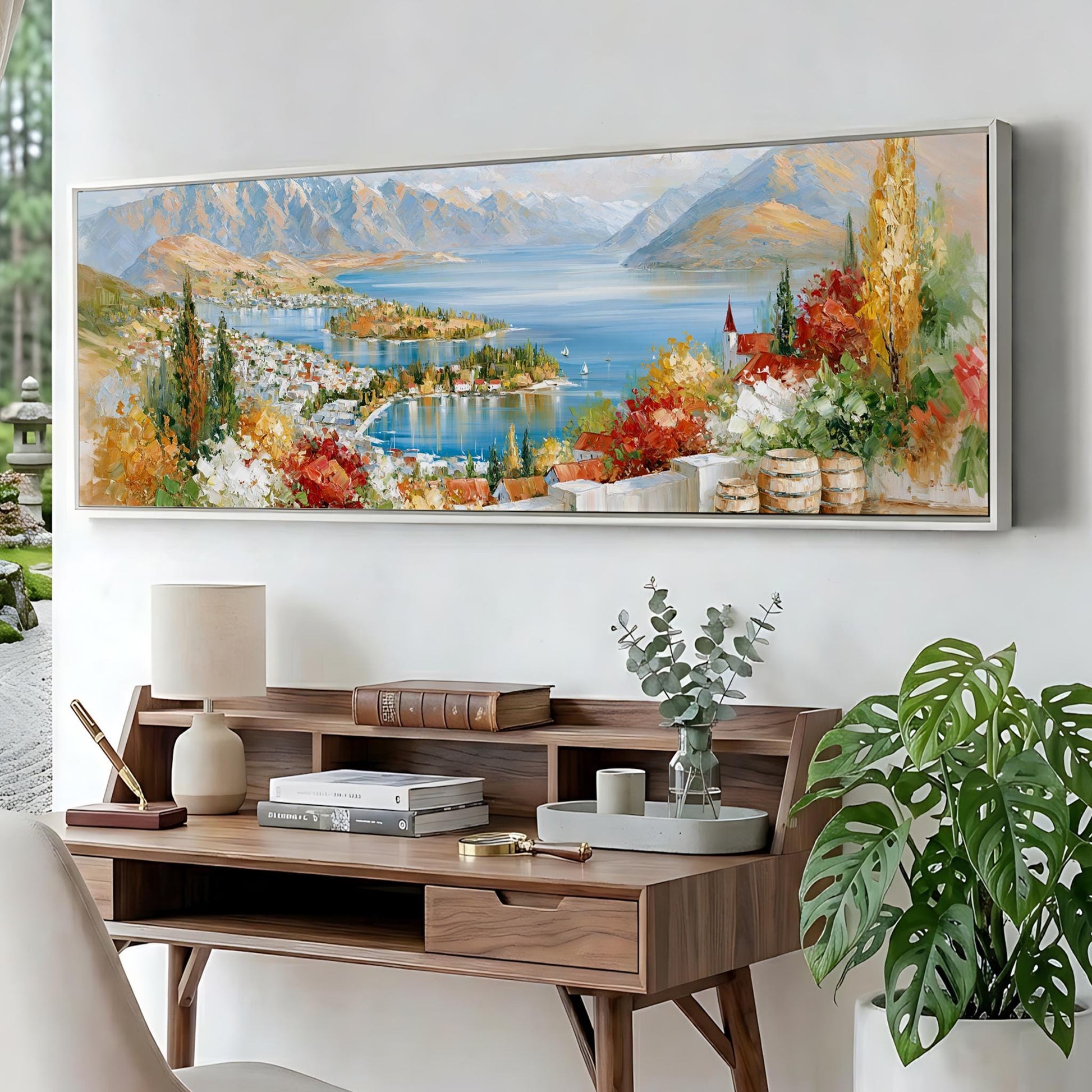 a large painting of a scenic landscape with mountains, a lake, and colorful foliage, hanging on a wall above a wooden desk with a lamp, books, and a vase of greenery.