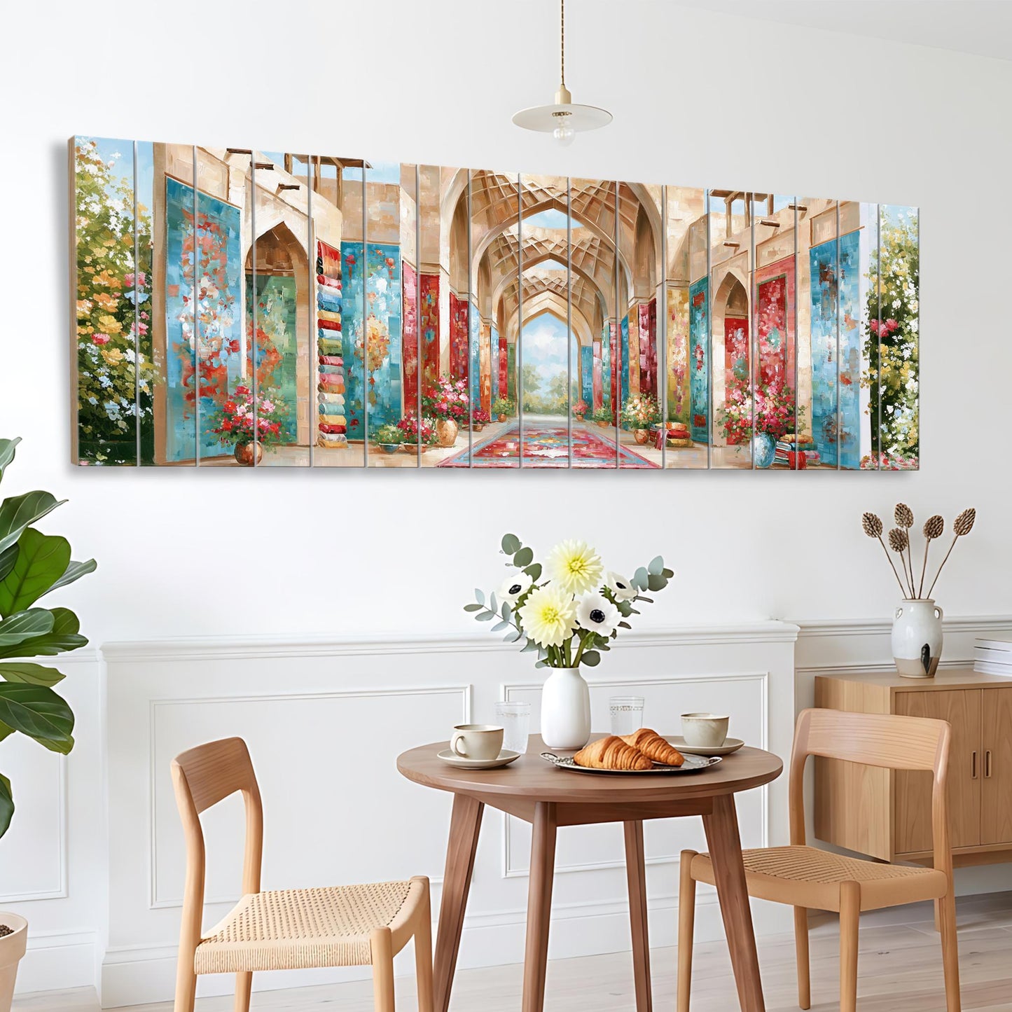 a dining area with a wooden table, two chairs, and a vase of flowers. Above the table, there is a large painting depicting an arched hallway with colorful walls and a chandelier.