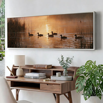 a large painting of ducks swimming on a lake, with a warm sunset in the background. The painting is hung on a wall above a wooden desk with a lamp, books, and a plant.