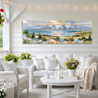 a cozy living room with a large, colorful landscape painting hanging on the wall. The painting features a serene mountain range and a tranquil lake, creating a peaceful and picturesque scene. The room is furnished with a white sofa, wicker chairs, and a round coffee table, all arranged around a white table with a vase of flowers. The overall atmosphere of the room is warm and inviting, with the natural elements of the painting complementing the comfortable furnishings and decor.