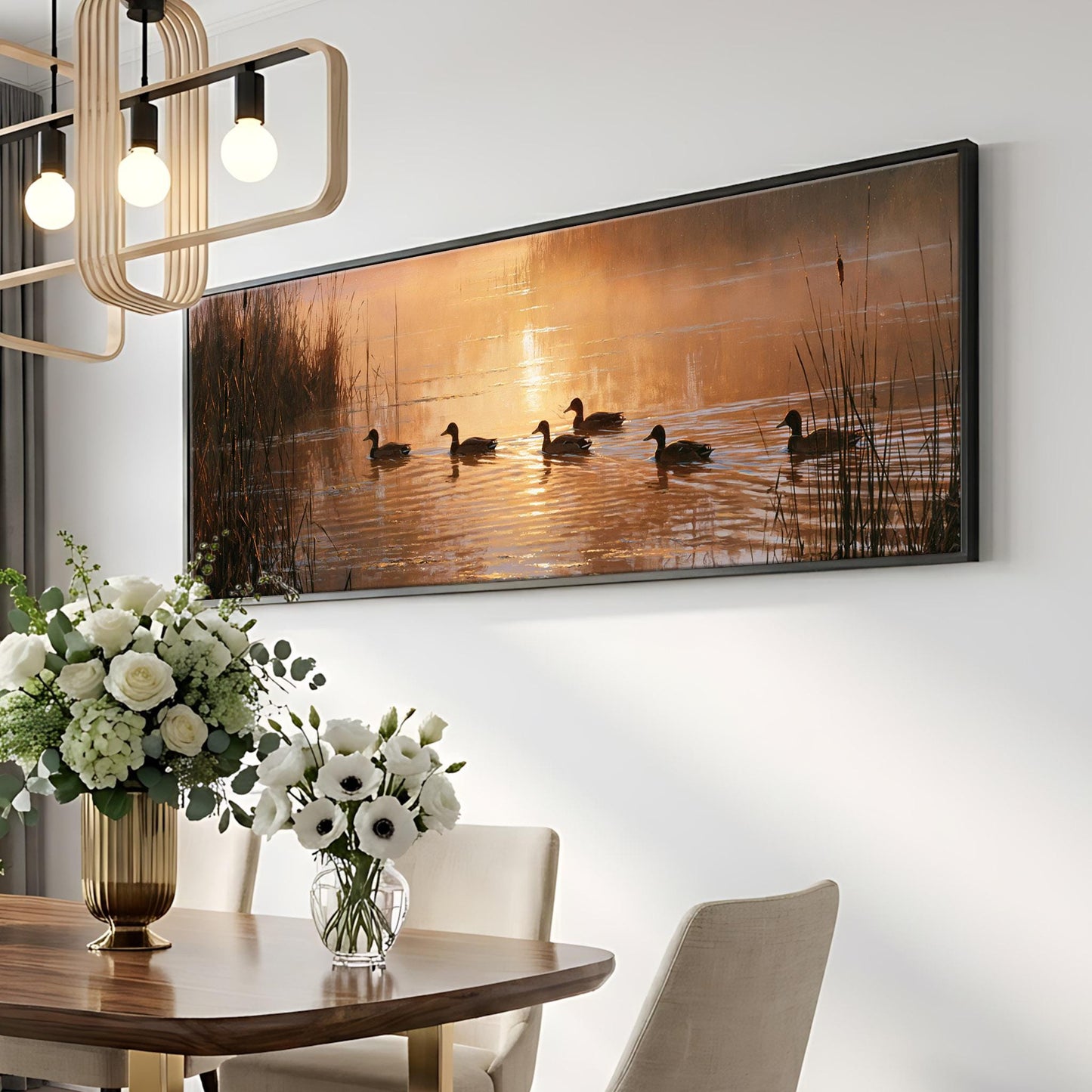 a large framed painting of ducks swimming on a lake, with a warm sunset in the background. The painting is hung on a wall above a dining table with a vase of white flowers on it.