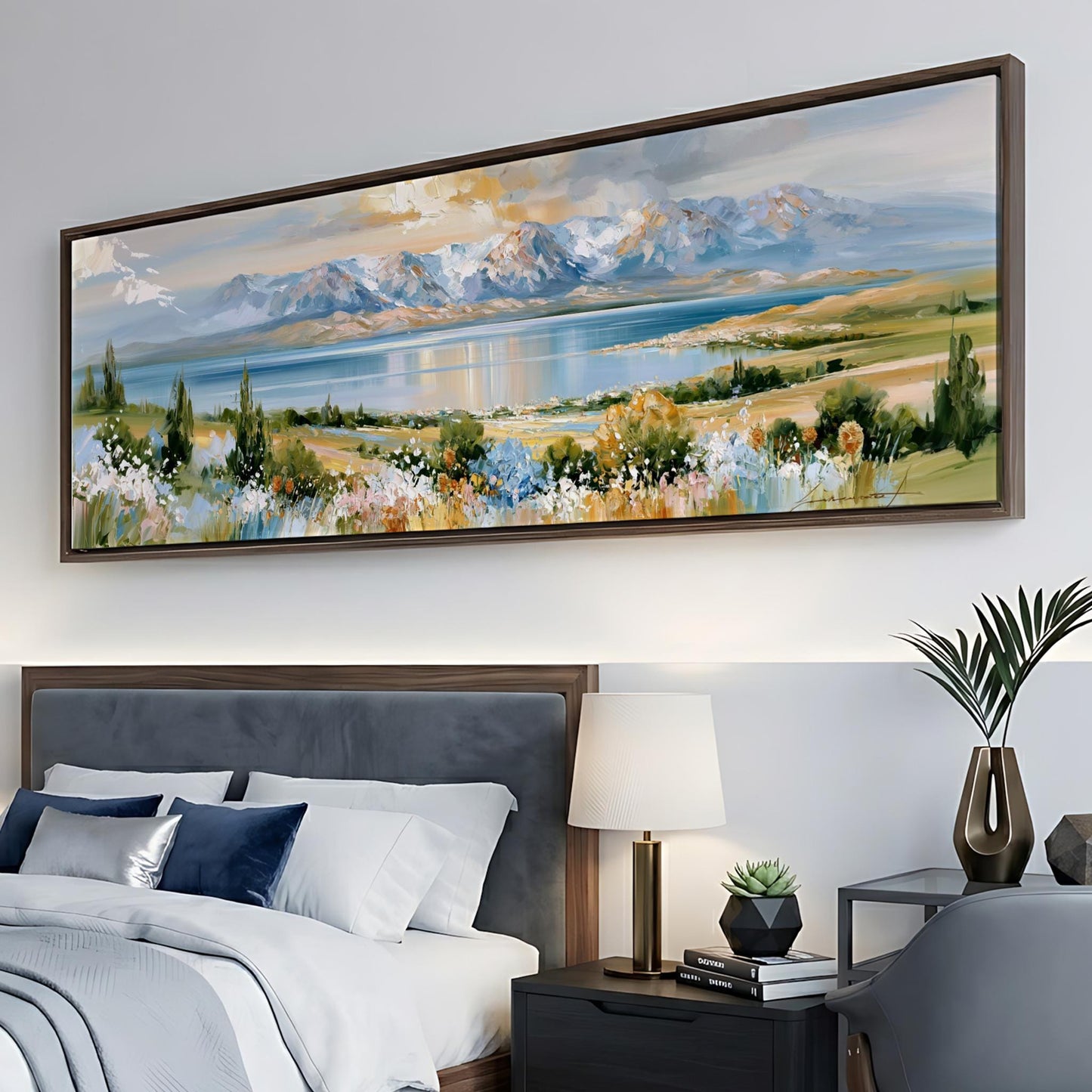 a large landscape painting hanging on a wall above a bed with a gray headboard. The painting depicts a serene mountain landscape with a lake and colorful flowers. The room also includes a nightstand with a lamp, a chair, and a plant, creating a cozy and inviting atmosphere.