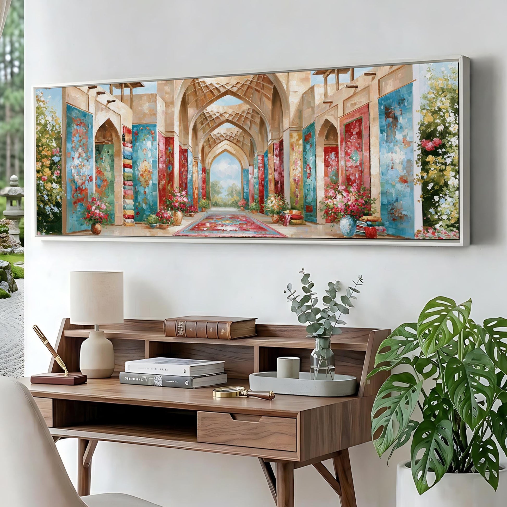 a large painting of an ornate hallway with arches and colorful walls, hanging on a wall above a wooden desk with a lamp, books, and a vase of flowers.