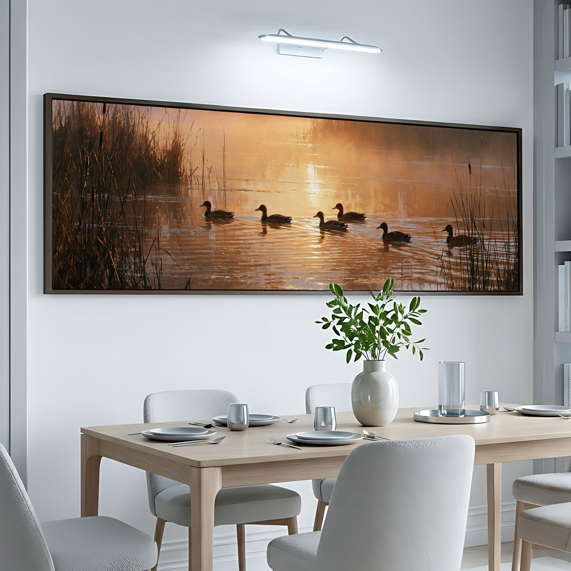 a large, framed painting of ducks swimming on a lake, with a warm sunset in the background. The painting is hung on a white wall above a dining table set with plates, silverware, and a vase of flowers.