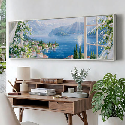 a large painting of a scenic landscape with a river, mountains, and a town, hanging on a wall above a wooden desk with a lamp, books, and a vase of flowers.