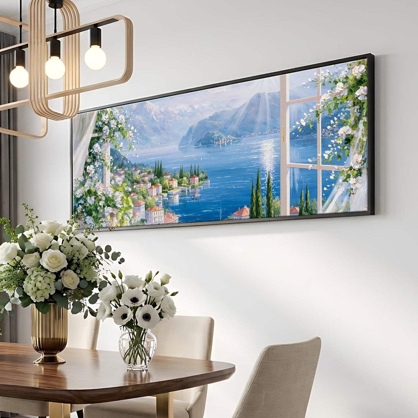 a large framed painting of a scenic view of a town and lake, hanging on a wall above a dining table with a vase of flowers.