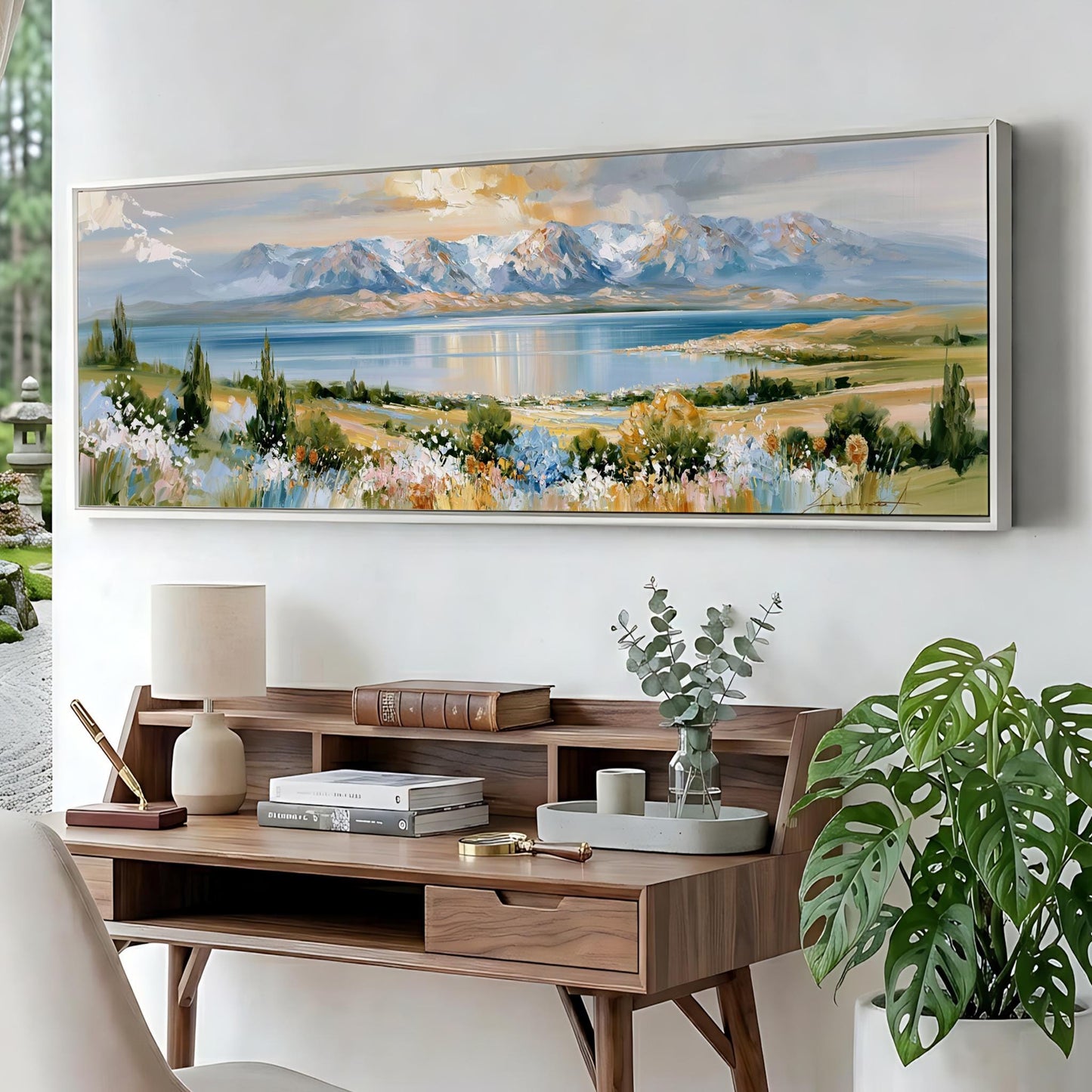 a large landscape painting hanging on a wall above a wooden desk with a lamp, books, and a vase of greenery.