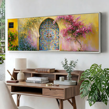 a large, colorful painting of a garden scene with a mosaic archway, a pink flowering tree, and a yellow wall. The painting is displayed on a wall above a wooden desk with a lamp, books, and a vase of flowers.