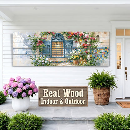 a large wooden sign that reads "Real Wood Indoor & Outdoor", with a painting of a house and flowers displayed on the wall above it. There are also potted plants and a wicker basket on the porch, adding to the overall aesthetic of the scene.