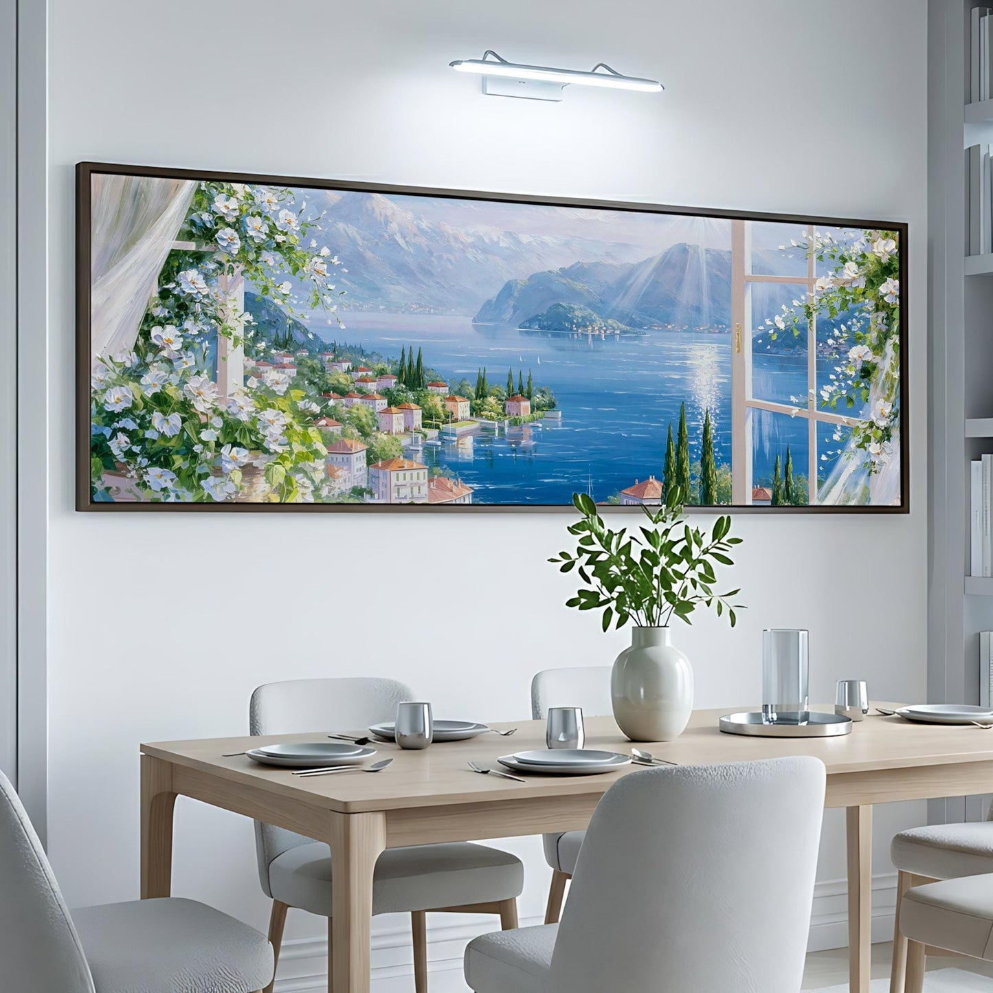 a large, framed painting of a scenic landscape with a river, mountains, and buildings, hanging on a white wall above a dining table with chairs.