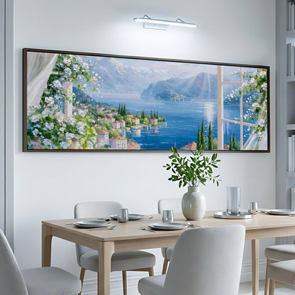 a large, framed painting of a scenic landscape with a river, mountains, and buildings, hanging on a white wall above a dining table with chairs.