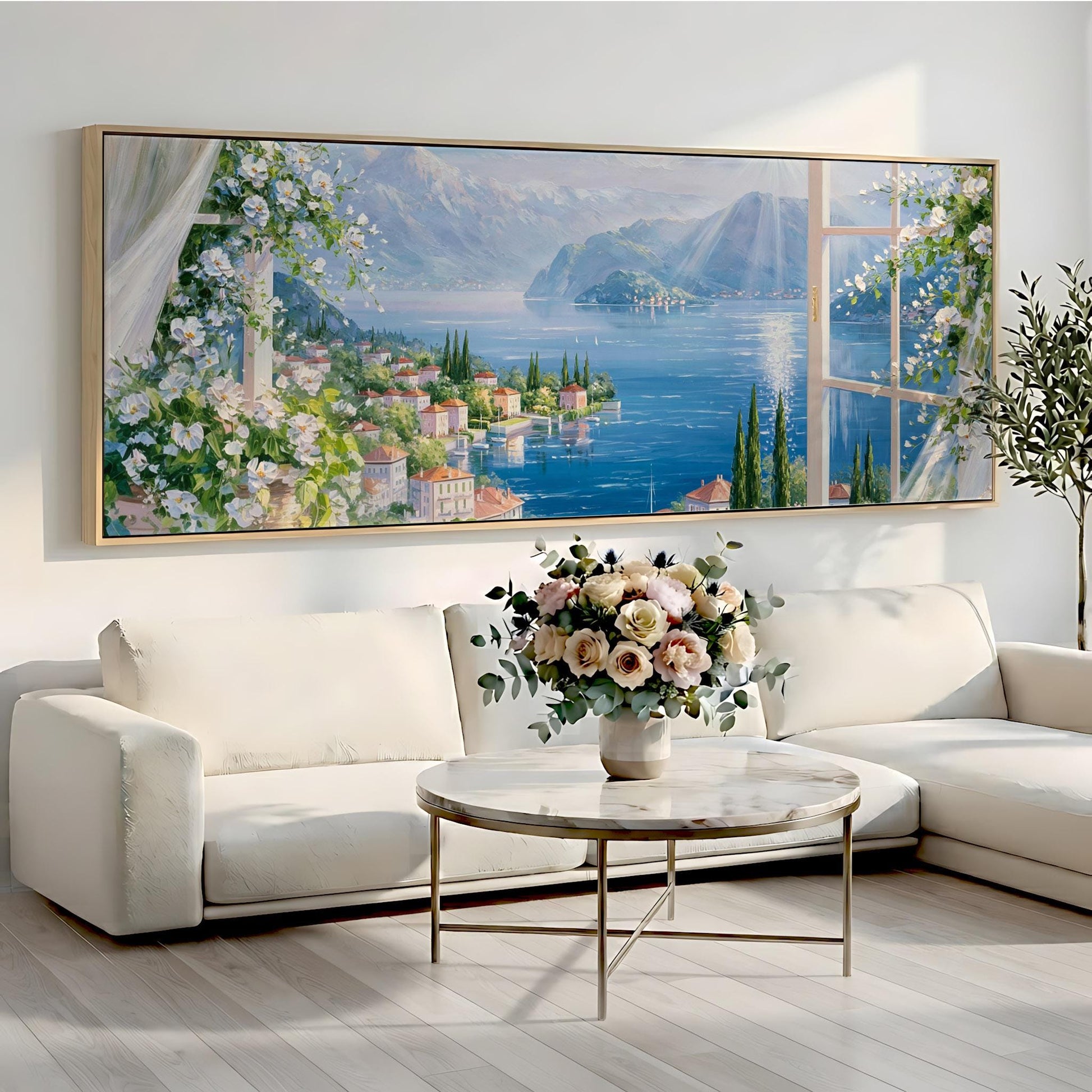 a large painting of a scenic landscape with a river, mountains, and a town, hanging on a wall above a white sofa and a round coffee table with a vase of flowers.