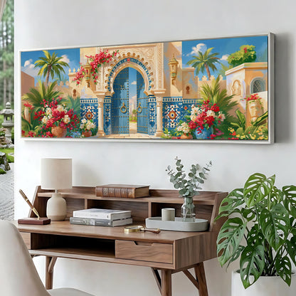a large, colorful painting of an ornate, blue and yellow gate with arches, surrounded by a garden with palm trees and flowers. The painting is displayed on a wall above a wooden desk with a lamp, books, and a vase of flowers.