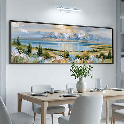 a large, framed landscape painting hanging on a white wall above a dining table with white chairs. The painting depicts a serene mountain landscape with a lake and colorful flowers in the foreground.