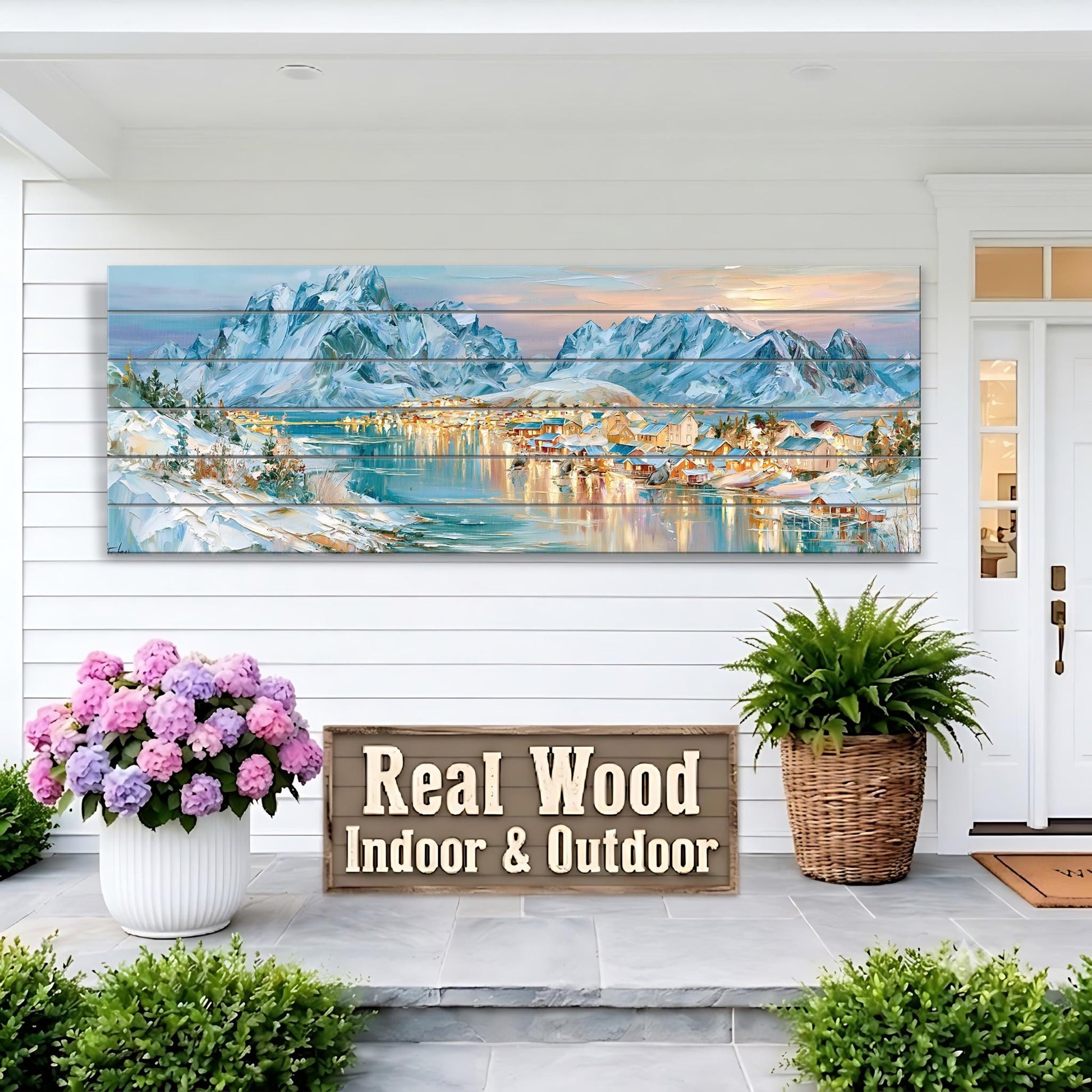 a large wooden panel hanging on a white wall, depicting a scenic mountain landscape with a town in the distance. The panel is mounted above a wooden sign that reads "Real Wood Indoor & Outdoor". The sign is placed on a porch, with two potted plants and a large bouquet of pink flowers nearby.