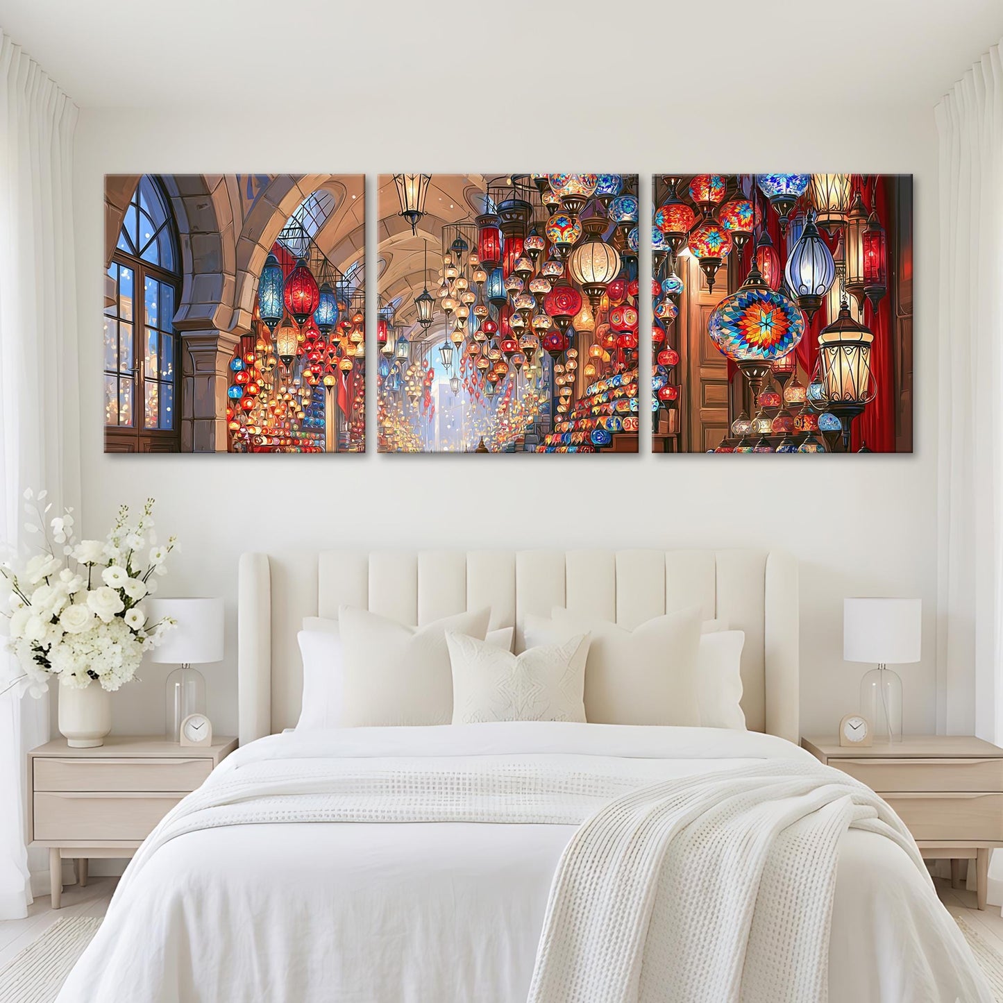 A large, white bed with a white comforter and pillows, flanked by two nightstands with lamps. Above the bed are three large, colorful paintings depicting a vibrant and ornate interior with hanging lanterns and lights.