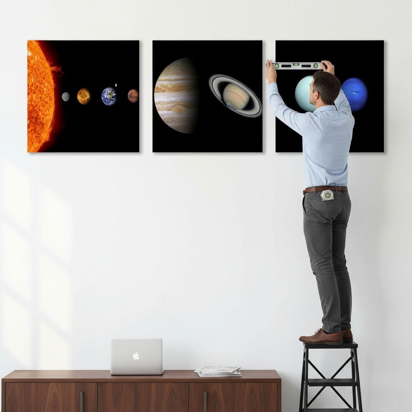 A man is standing on a ladder and hanging three framed images of planets on a wall.