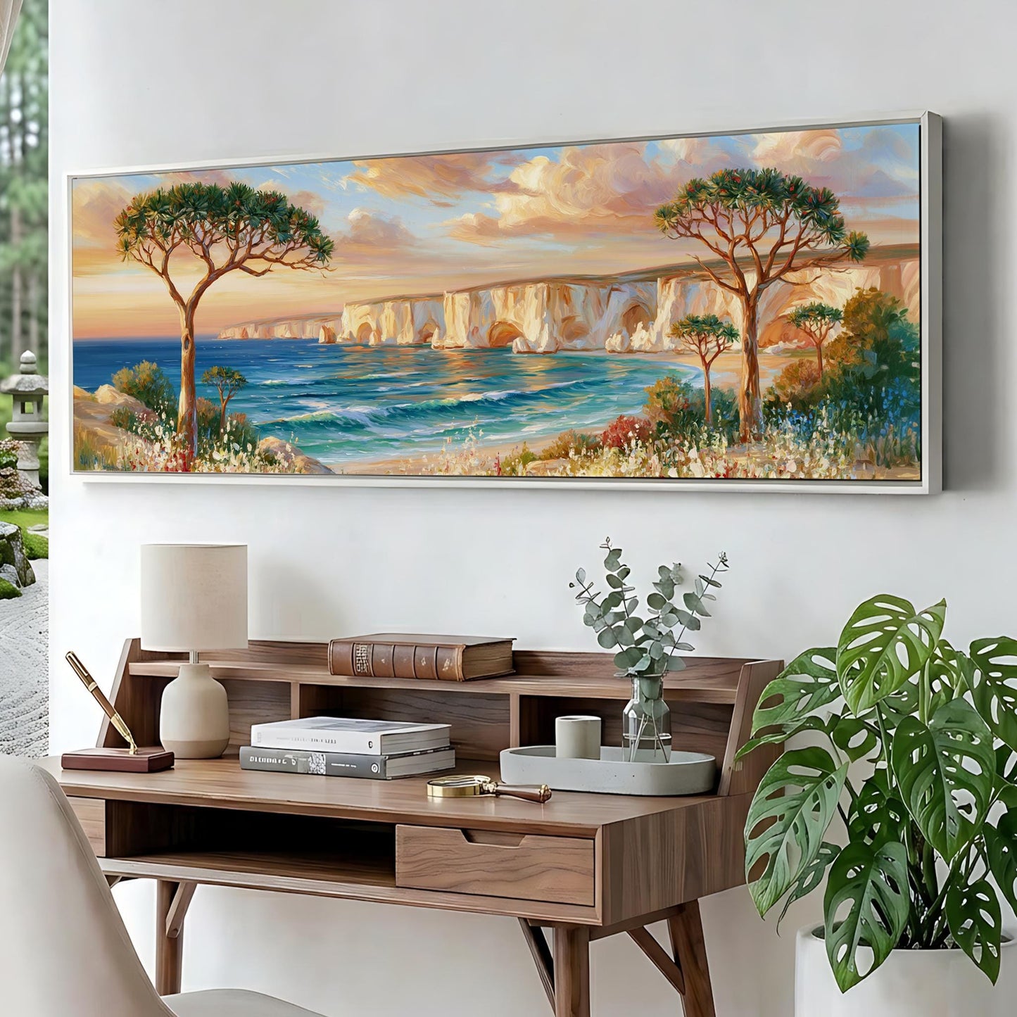 a large painting of a serene landscape with a beach, trees, and a body of water, hanging on a wall above a wooden desk with a lamp, books, and a vase of greenery.