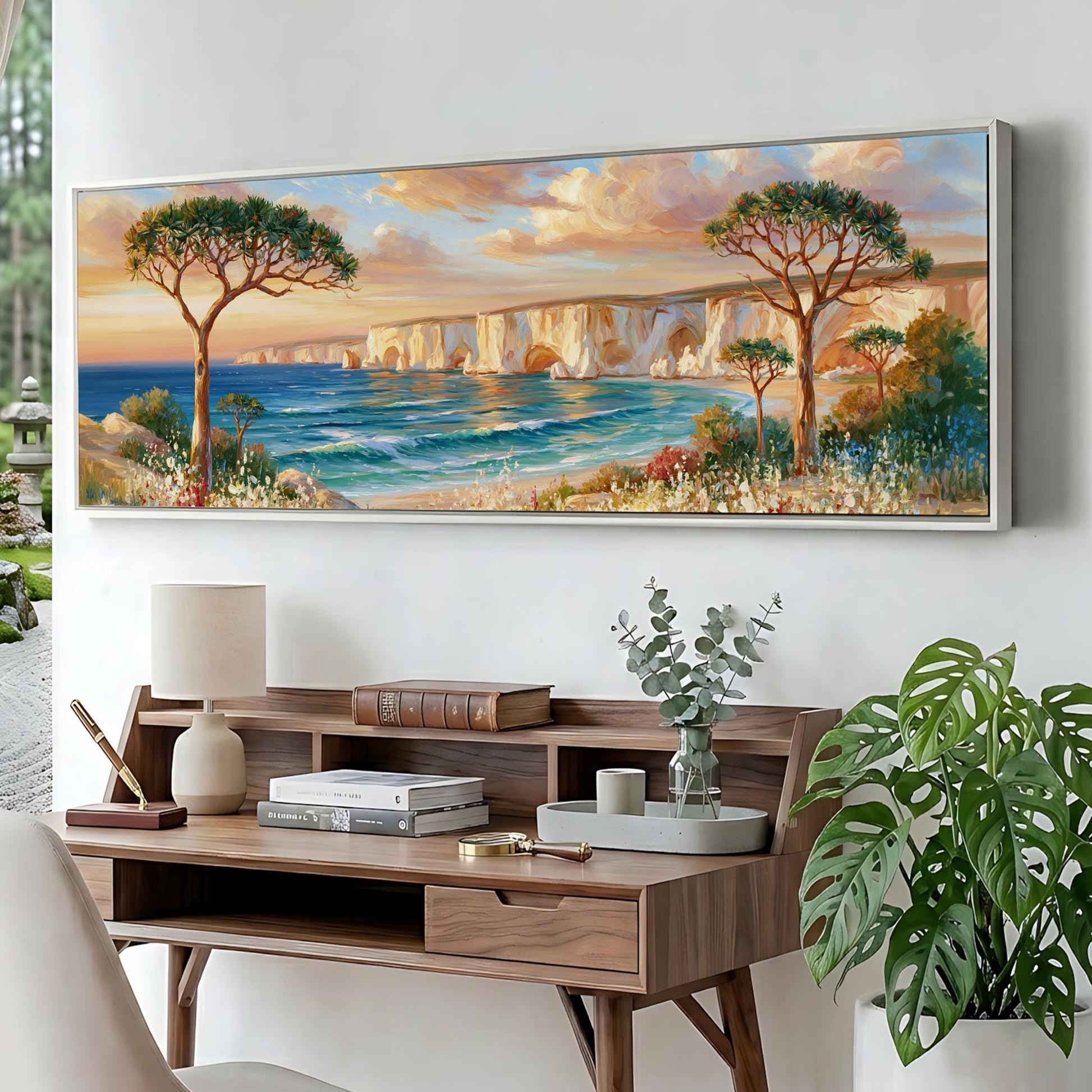 a large painting of a serene landscape with a beach, trees, and a body of water, hanging on a wall above a wooden desk with a lamp, books, and a vase of greenery.