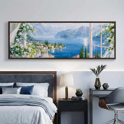a large painting of a scenic view of a coastal town with mountains in the background, hanging on a wall above a bed with a gray headboard.