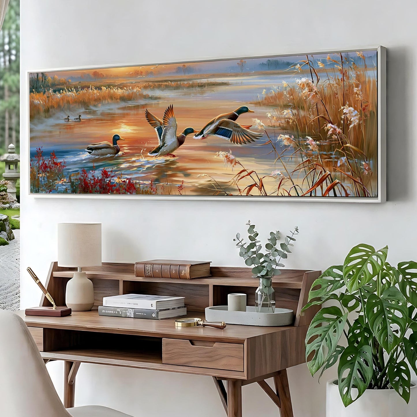 a large painting of ducks swimming in a pond, hanging on a wall above a wooden desk with various items on it.